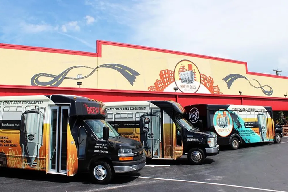 Three buses with custom beer-themed wraps are parked in front of a building with a large Brew Bus sign and beer-related graphics
