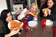 Three people are joyfully eating large slices of pizza at an outdoor table.