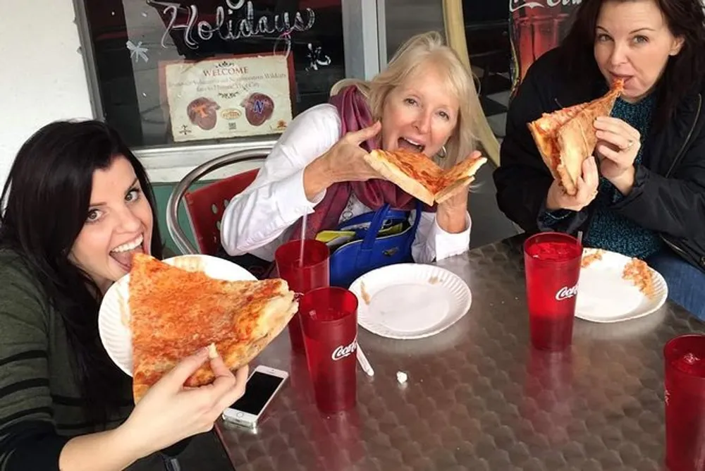 Three people are joyfully eating large slices of pizza at an outdoor table