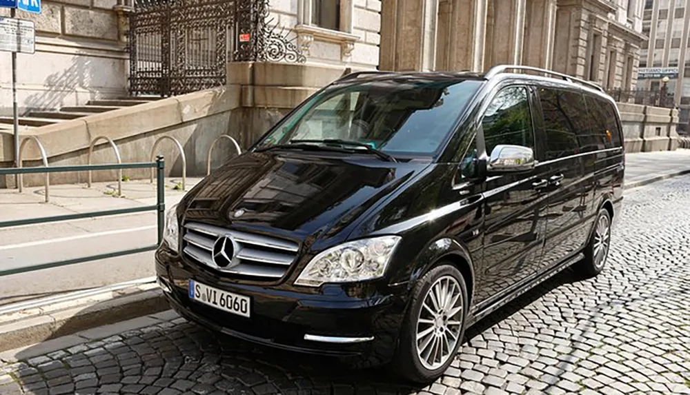 A black Mercedes-Benz van is parked on a cobblestone street next to a sidewalk with metal railings reflecting the surrounding buildings