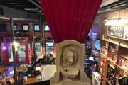 The image shows an indoor setting with a large stone fireplace featuring a lion's head, viewed from an elevated position behind a red curtain, overlooking a room with a bar and seating areas, all with a warm and cozy ambiance.