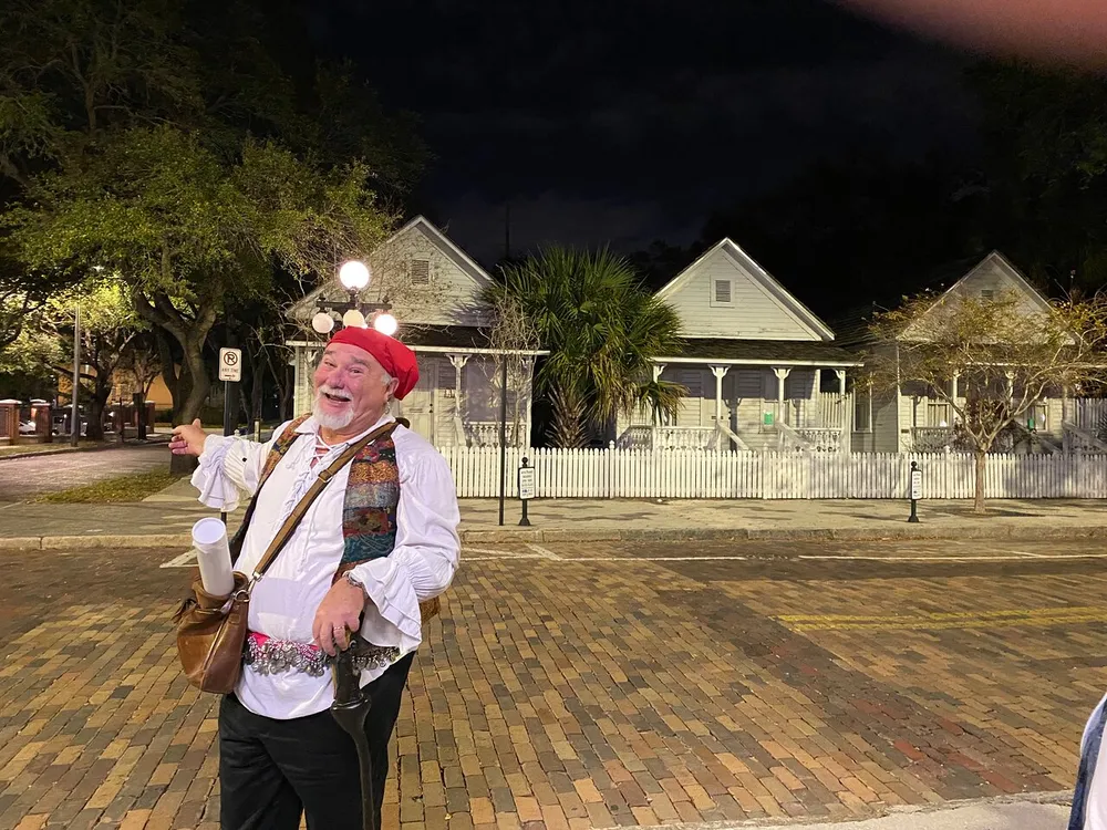 A jovial man in eclectic attire gestures with a smile on a street with historic-looking houses in the background at night