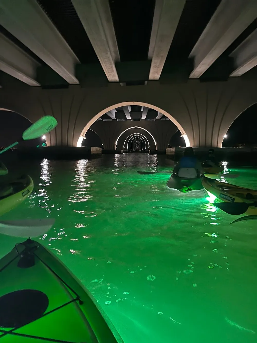 Kayakers paddle through a series of illuminated archways under a bridge at night with green lights casting a glow on the water beneath them