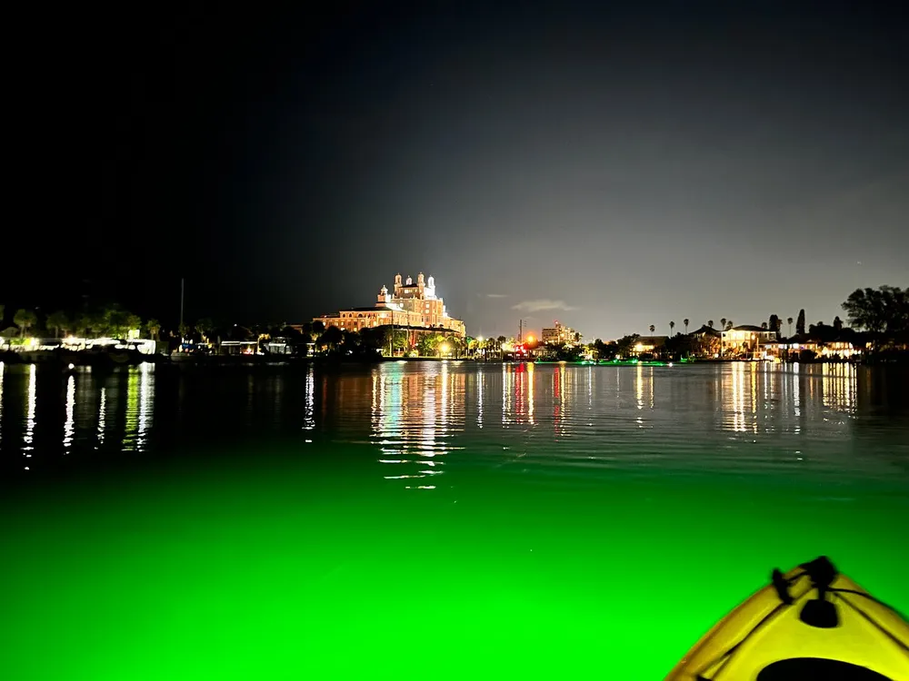 This image depicts a nighttime view from a kayak on a body of water showcasing a lit-up waterfront with buildings and reflections of lights on the water
