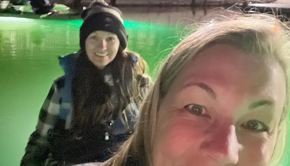 The image shows a close-up of a persons face in the foreground with another smiling person positioned in the background both wearing warm clothing suggesting a cold environment
