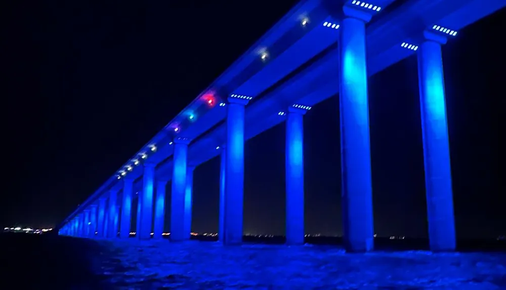 A bridge is illuminated with blue lights at night creating a striking contrast with the dark sky and water below