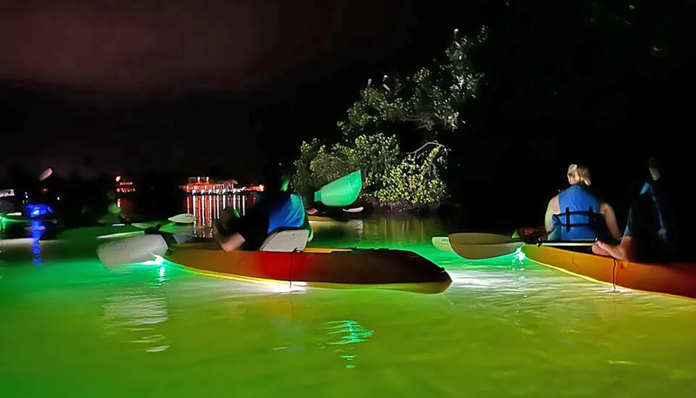 A group of people are night kayaking with the water illuminated by underwater lights attached to their kayaks creating a serene glowing effect