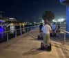 A group of people are riding Segways along a waterfront promenade at dusk with city lights reflecting on the water