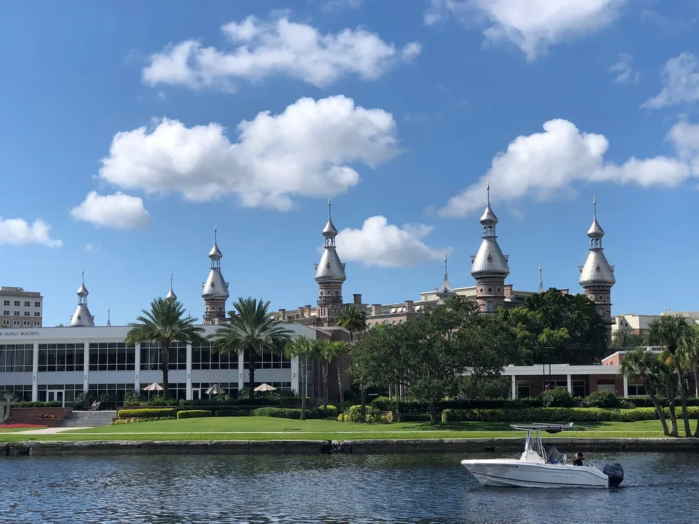 The image shows a boat on the water in front of a picturesque building with distinctive spired rooftops palm trees and a clear blue sky with scattered clouds