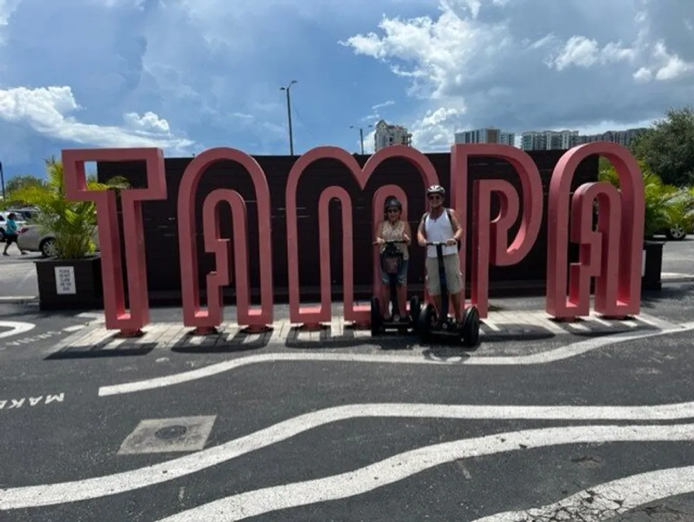 Two people on Segways are posing in front of a large red sign that spells out TAMPA