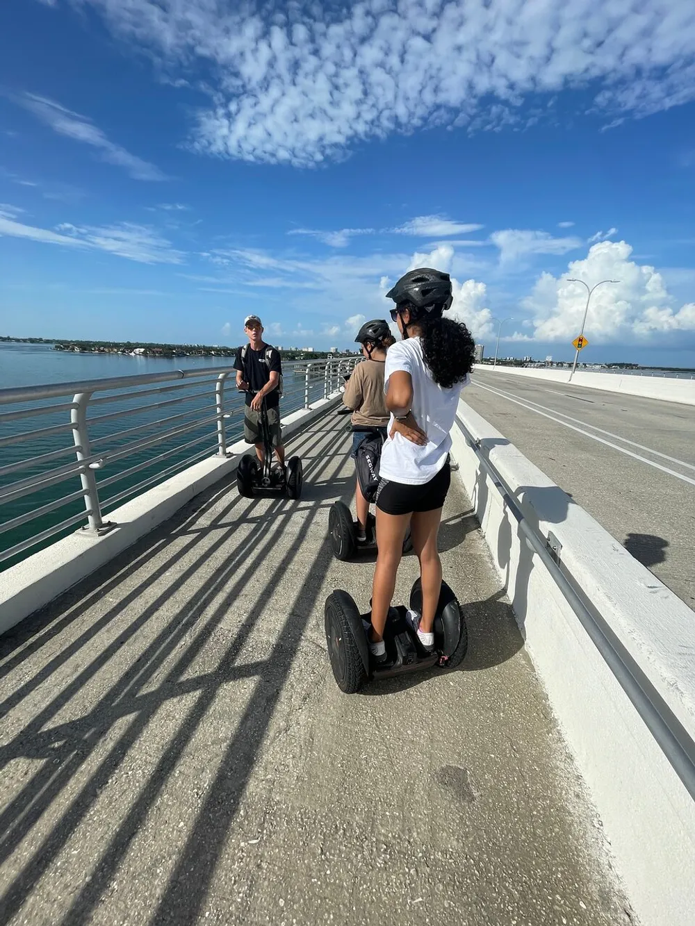 Three people are riding Segways on a sunny bridge over water with a clear blue sky above