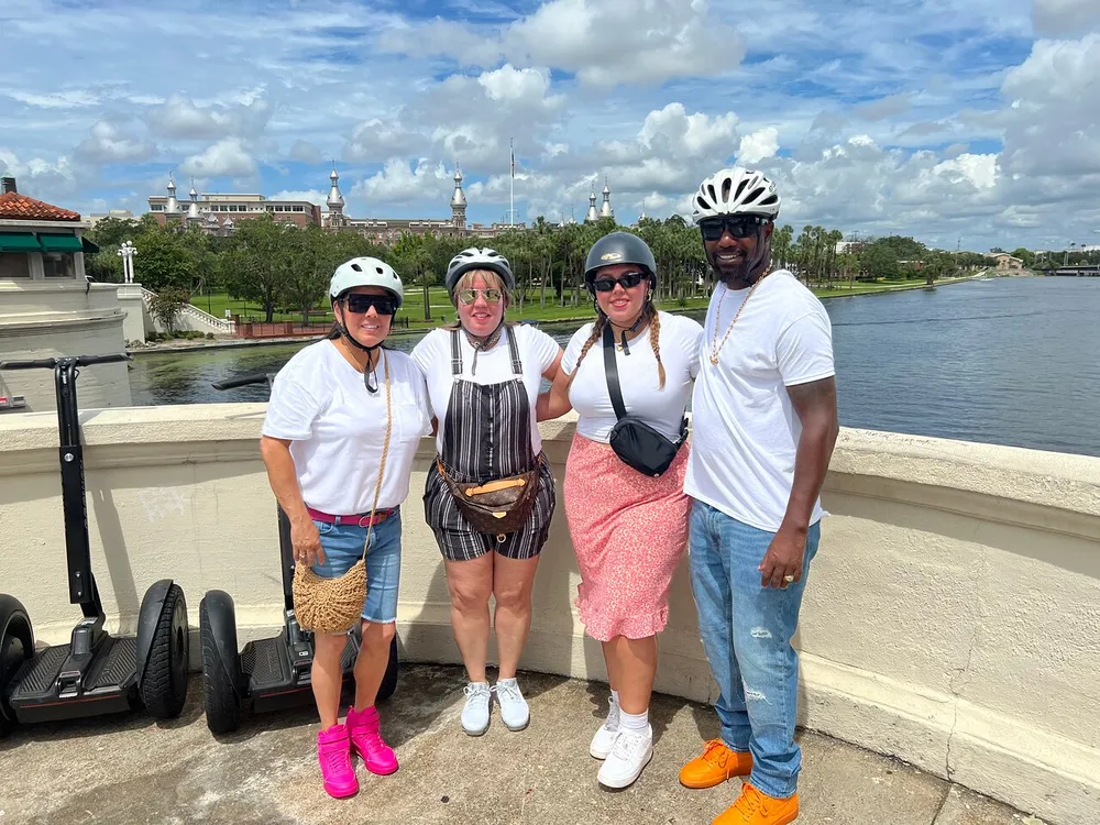 Four people wearing helmets and casual attire are posing for a photo beside a scenic riverfront with two electric scooters parked to their left