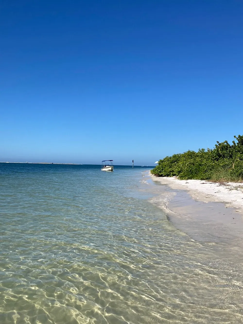 The image shows a serene beach scene with clear turquoise waters a stretch of white sand green vegetation to the side and a solitary boat floating near the shore under a vast blue sky