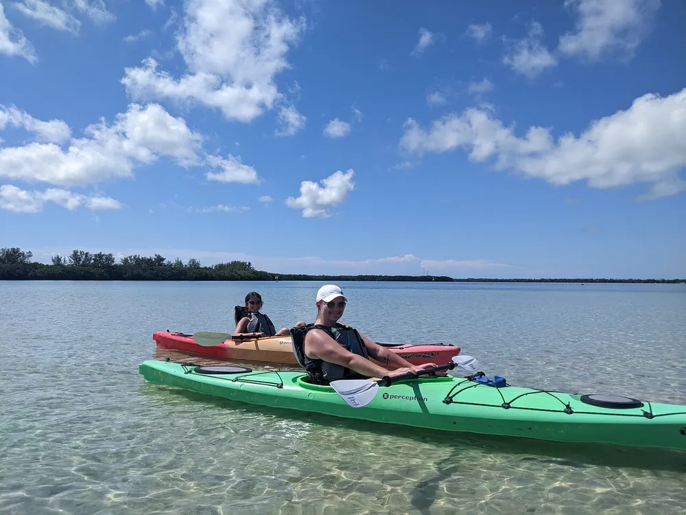 Two people are kayaking on a calm clear water body with a backdrop of a blue sky dotted with clouds