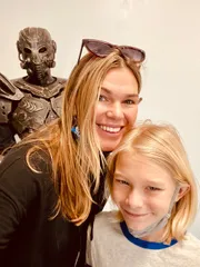A smiling woman and a child pose for a selfie with a person wearing a robot-like costume in the background.