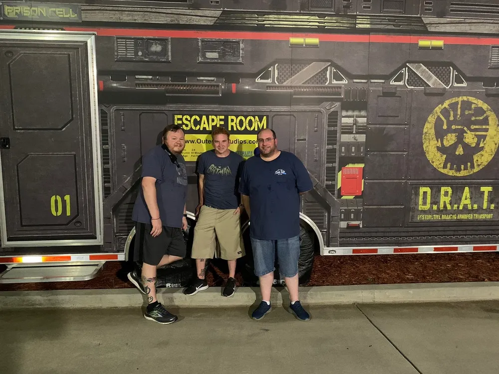 Three individuals are posing for a photo in front of a backdrop that resembles a sci-fi themed escape room entrance