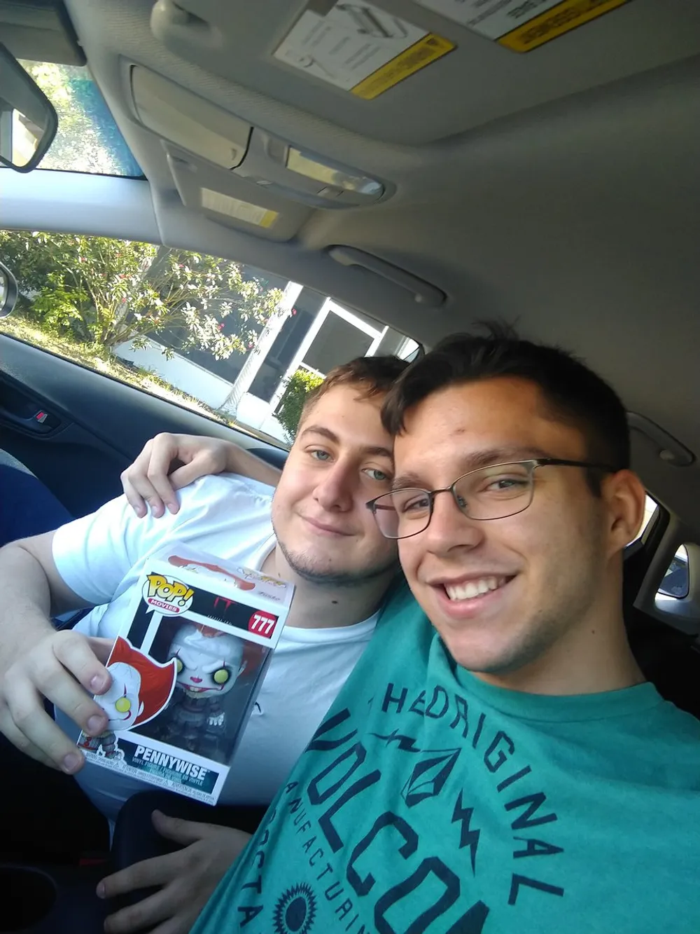 Two smiling individuals are taking a selfie in a car with one of them holding a Funko Pop figure of the character Pennywise from It