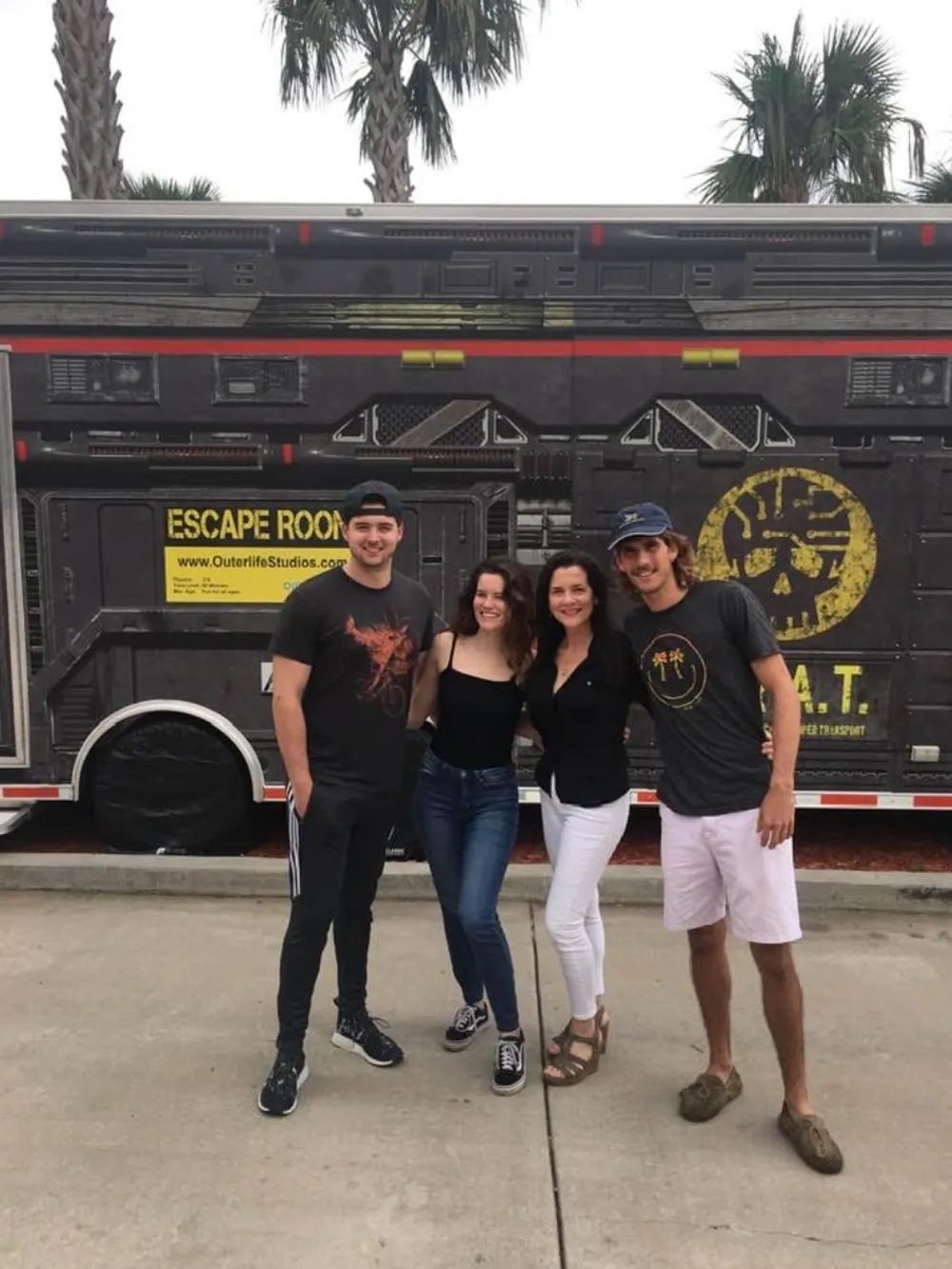 Four people are smiling and posing in front of a vehicle themed as an Escape Room from Outerlife Studios with palm trees in the background