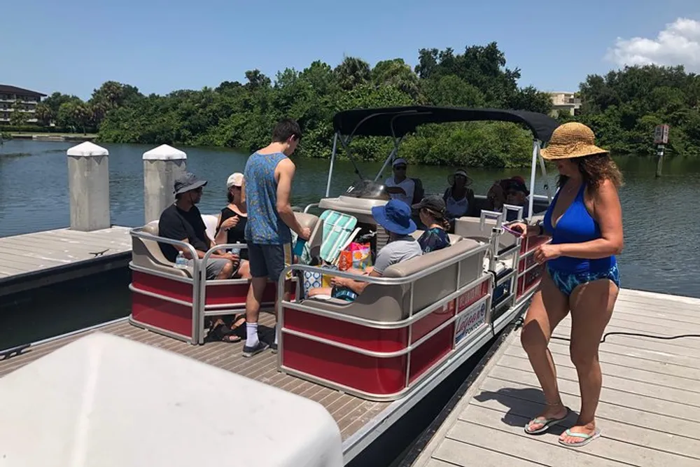 A group of people are preparing for a day on the water with one person standing on the dock next to a pontoon boat filled with supplies and passengers
