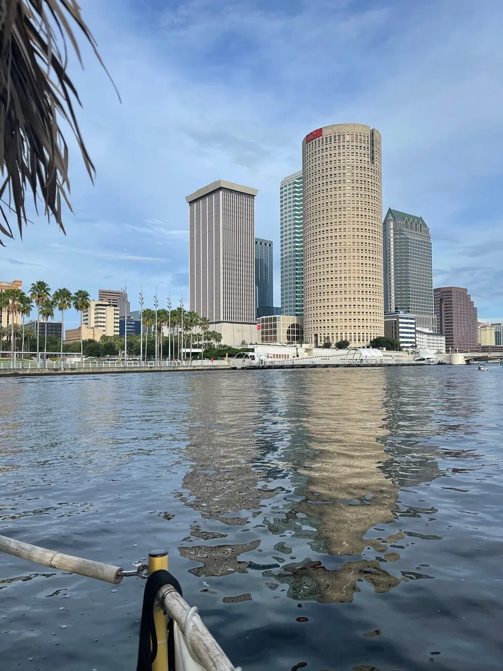 The image shows a waterfront view of a city skyline with towering skyscrapers and their reflections in the calm water taken from behind a partial palm frond and a boats railing