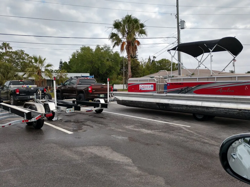 A red pontoon boat named Avalon is being towed by a brown pickup truck in a parking lot with other vehicles and trailers nearby under a cloudy sky with palm trees in the background