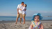 A baby wearing a large sunhat sits in the foreground on a beach, observing the camera with a puzzled expression, while in the background a couple embraces with a kiss.