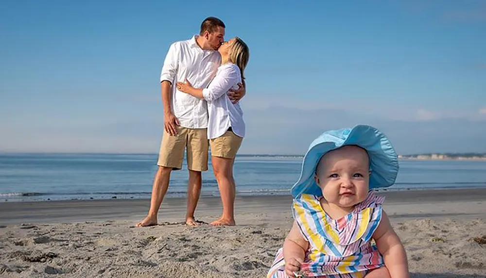A baby wearing a large sunhat sits in the foreground on a beach observing the camera with a puzzled expression while in the background a couple embraces with a kiss