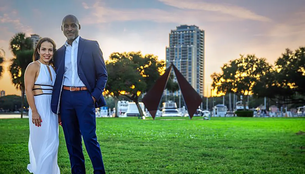 A couple is posing for a photo on a grassy area with a scenic sunset and urban skyline in the background