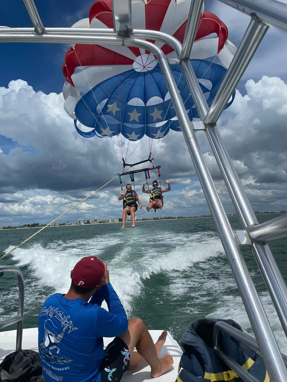 Two people are parasailing over the ocean tethered to a boat where an operator is observing them against a backdrop of clouds and a distant shoreline
