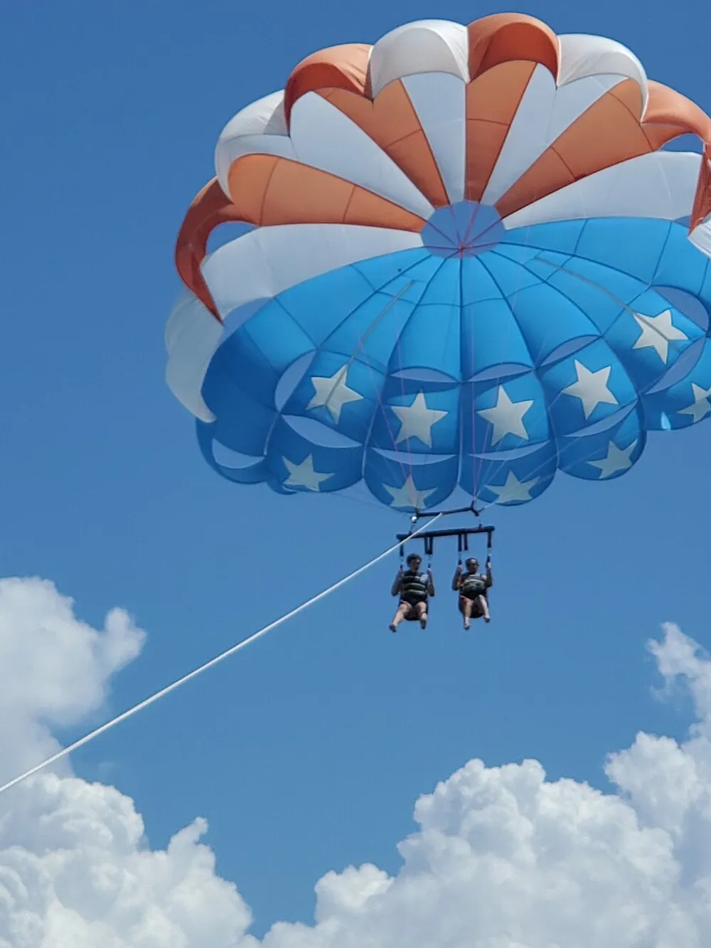 Two individuals are parasailing with a parachute adorned with stars and striped patterns high above the clouds against a clear blue sky