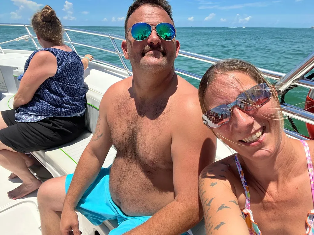 A couple takes a sunny selfie on a boat with a woman sitting in the background all enjoying a day at sea