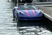 A blue sports car with pink striping is submerged in water next to a dock, appearing to have accidentally driven into the water.