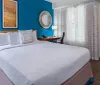 This image shows a neatly arranged hotel room with a large bed vibrant blue accent wall decorative art a work desk and a window dressed with sheer curtains
