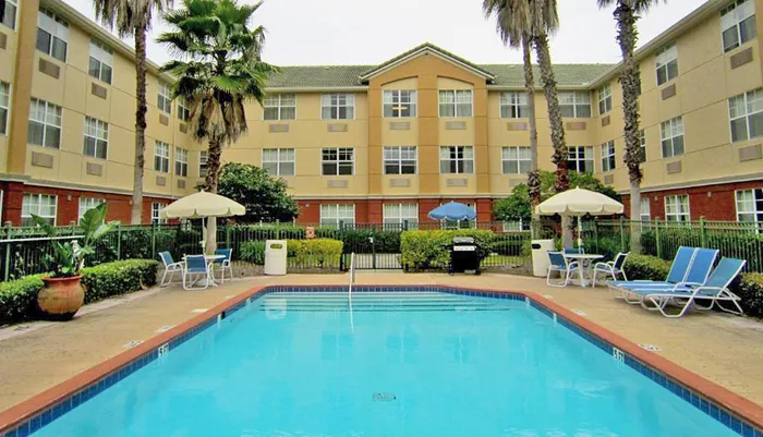 The image shows a swimming pool with lounge chairs and umbrellas in the courtyard of a multi-story hotel or apartment complex flanked by palm trees