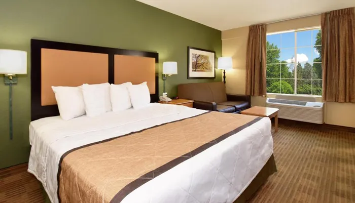 This image shows a neatly arranged hotel room with a large bed a leather couch bedside lamps a framed picture above the couch and a window with a view of trees