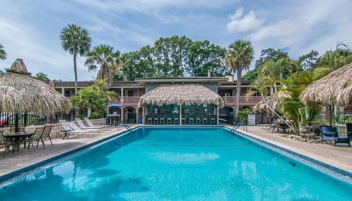 This image shows a large outdoor swimming pool with lounge chairs and thatched-roof structures flanked by palm trees and a two-story building with balconies in the background