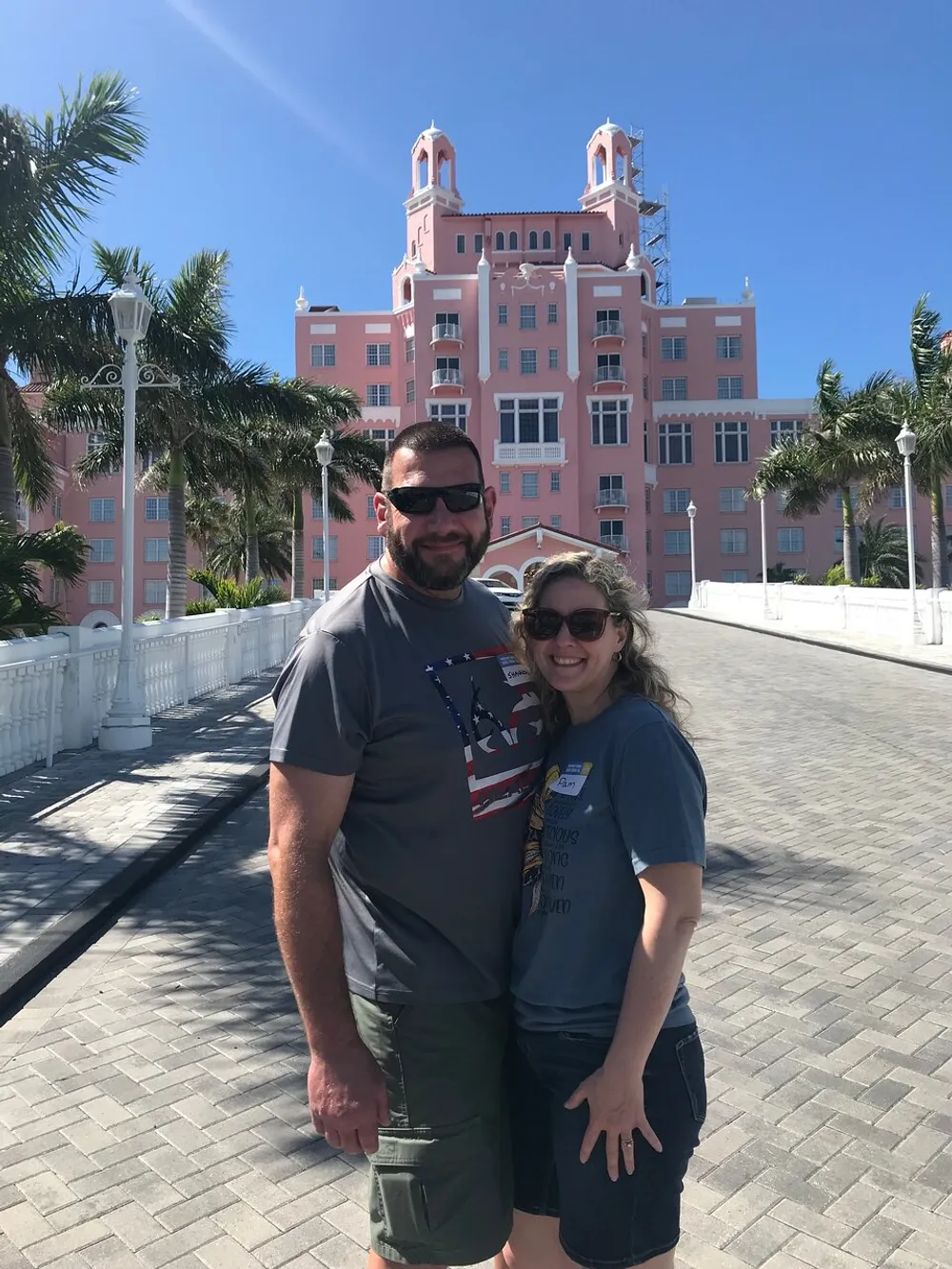 A smiling couple poses for a photo in front of a large pink building with palm trees around them under a clear blue sky