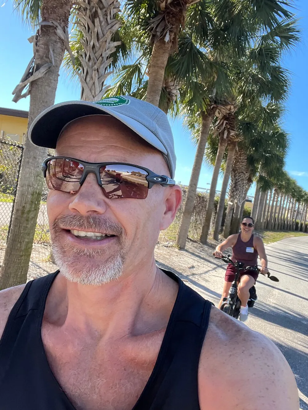 A person is taking a selfie with another individual riding a bike in the background both enjoying a sunny day outdoors lined with palm trees
