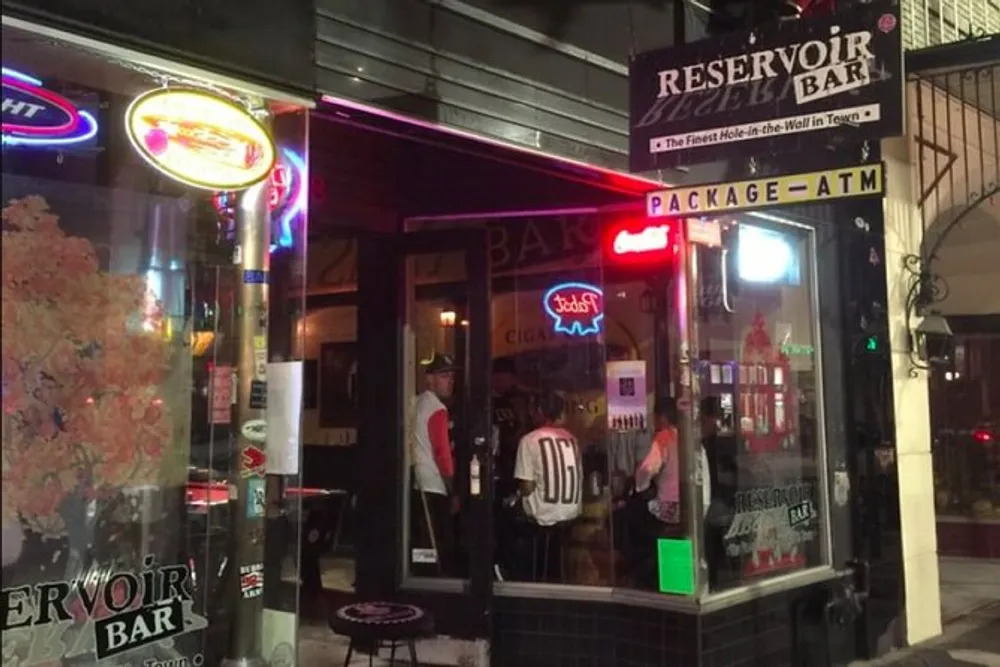The image shows a lively street view at night with a brightly neon-signed bar called Reservoir Bar and people visible inside