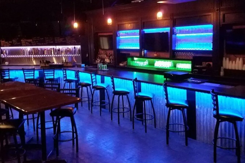 A dimly lit bar with blue accent lighting features an array of stools and a well-stocked counter with taps and illuminated shelves