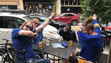 A group of people in casual attire and sunglasses are seated at an outdoor table, some with raised arms, exuding an energetic and carefree vibe.