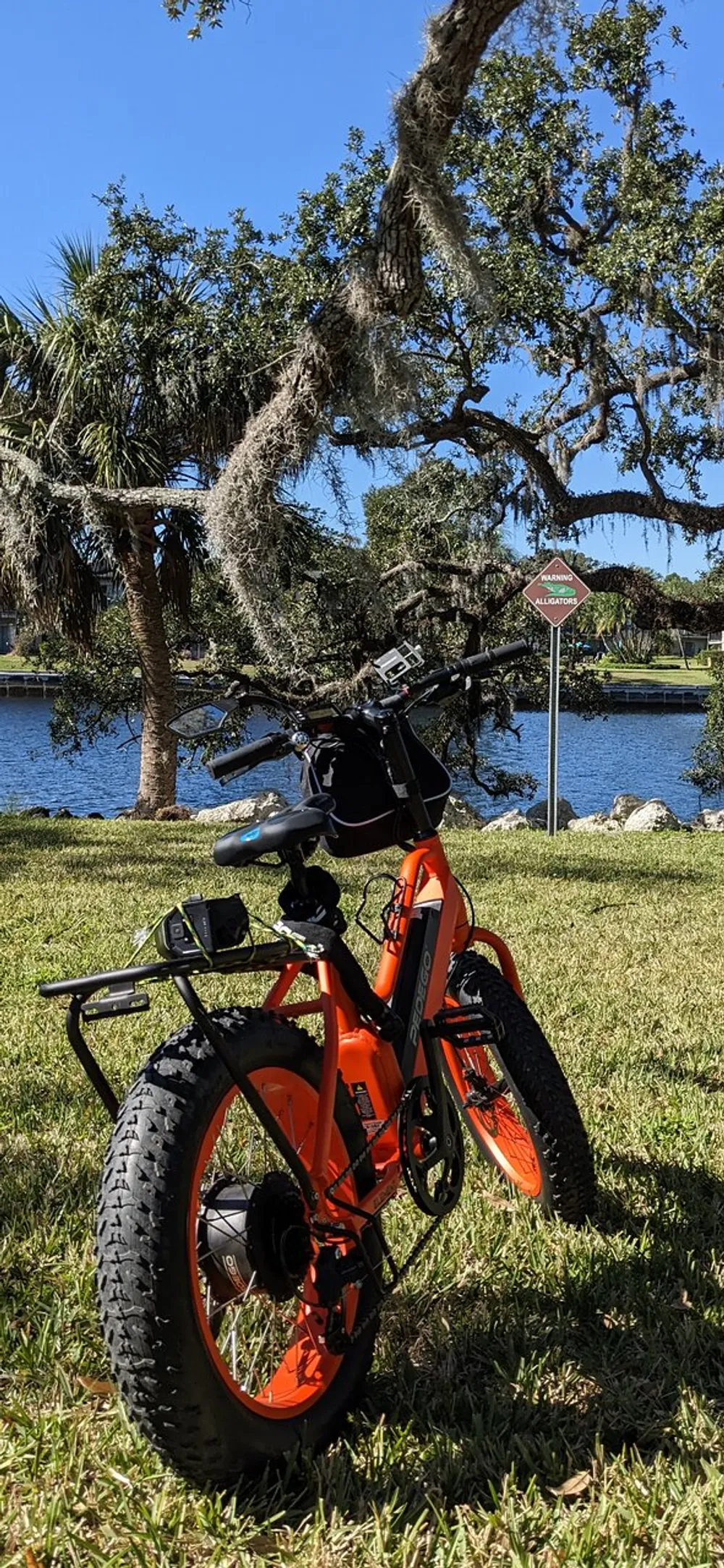 An orange electric bike with fat tires is parked on grass near a body of water with a Warning Alligators sign and Spanish moss-draped trees in the background