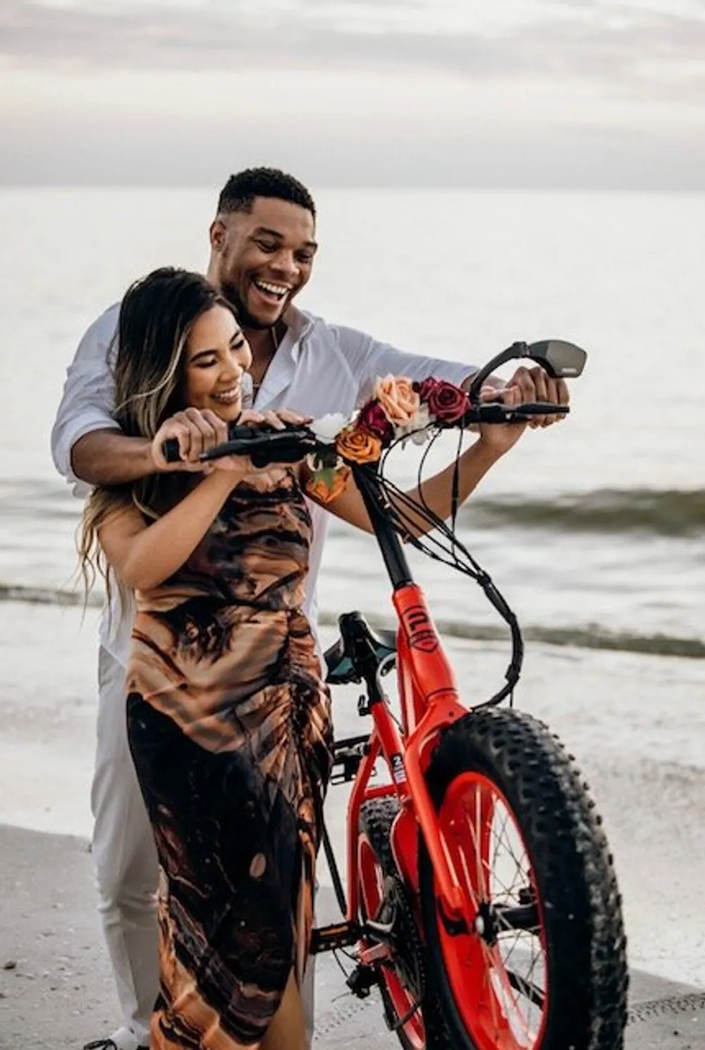 A smiling couple stands close together on a beach with the woman reaching out to touch the handlebar of a red fat-tire bicycle adorned with roses conveying a moment of joy and intimacy
