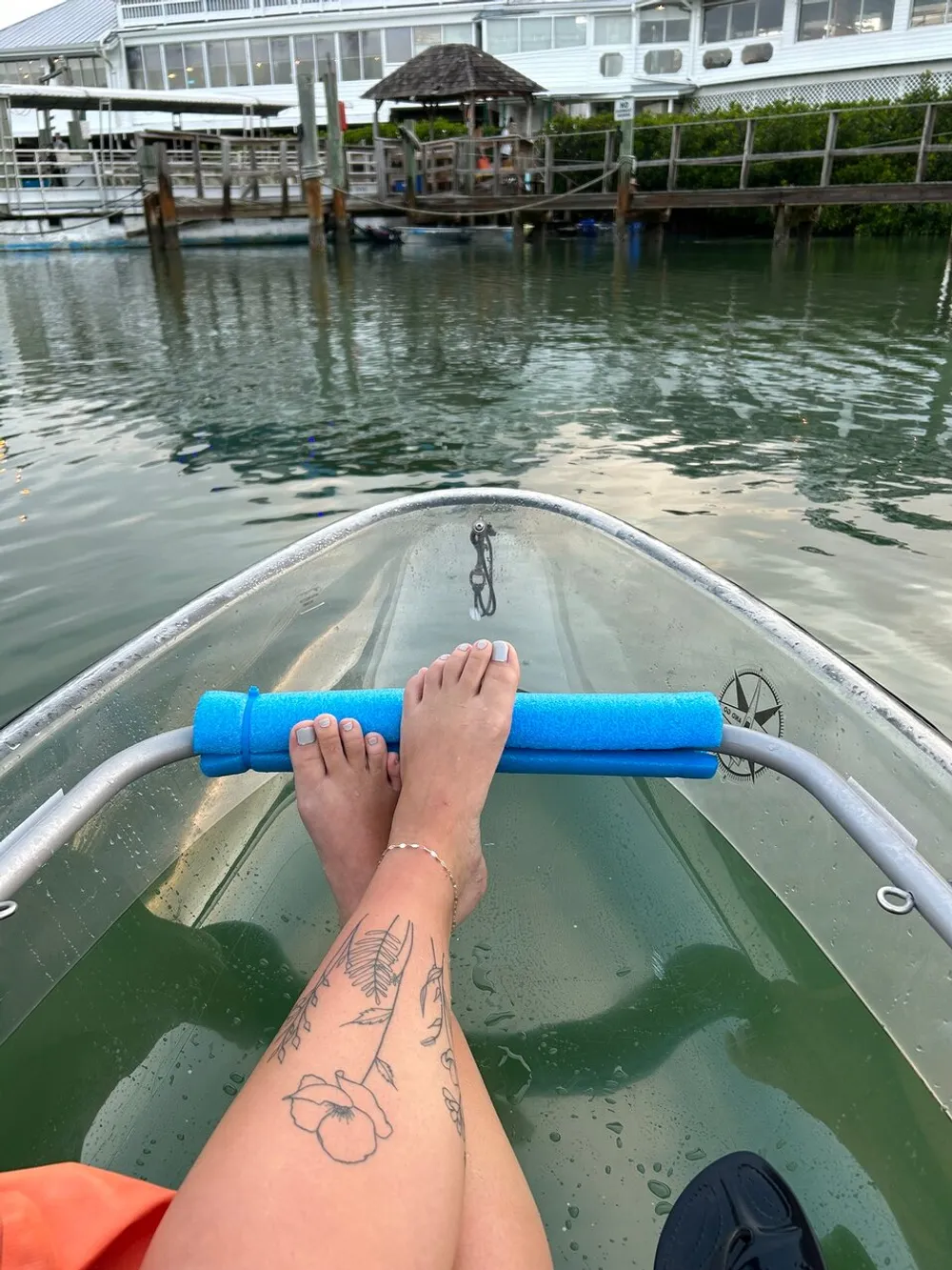 The image shows a persons bare feet resting on a blue foam pool noodle across a kayak with a scenic waterfront and a boat dock in the background