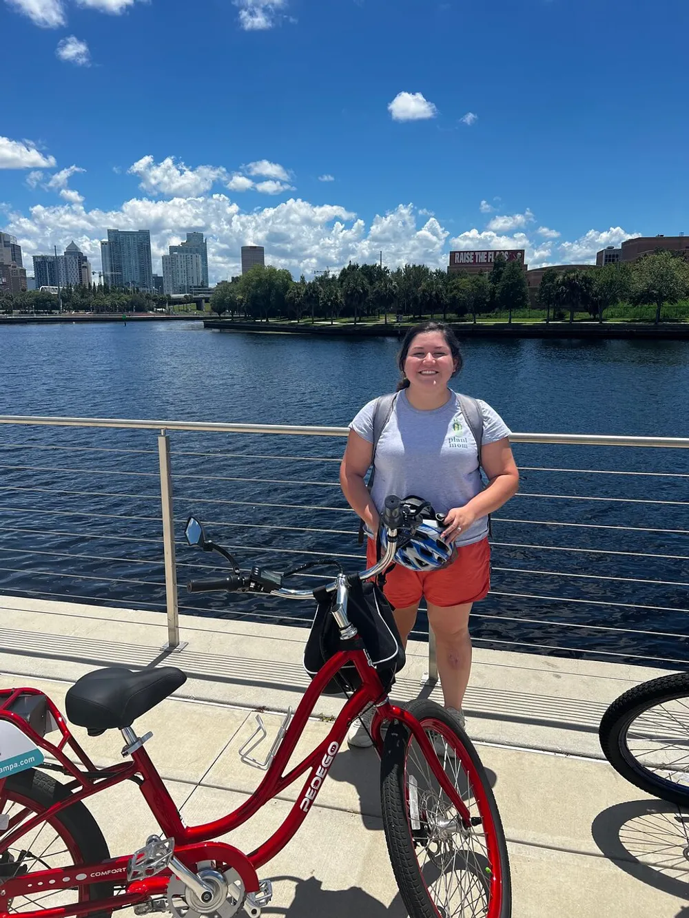 A person stands smiling next to a red bicycle on a sunny day with a cityscape and water in the background
