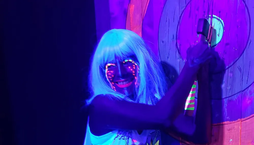 A person with neon face paint and a blue wig is smiling and posing under blacklight