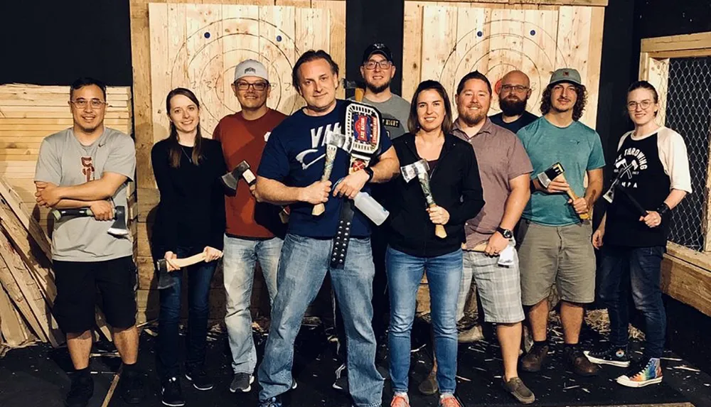 A group of people are posing with axes in front of wooden targets likely at an axe throwing facility