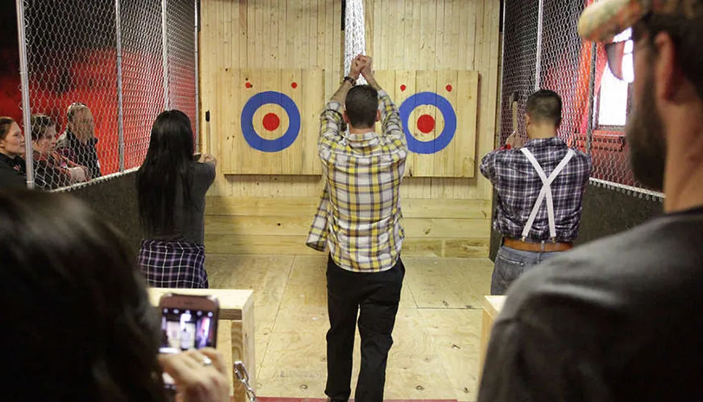 People are engaged in axe throwing at a target in an indoor facility with an onlooker capturing the moment on their smartphone
