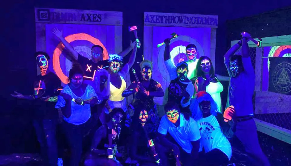 A group of people with neon face paint pose excitedly in a glow-in-the-dark setting holding axes and standing in front of axe throwing targets