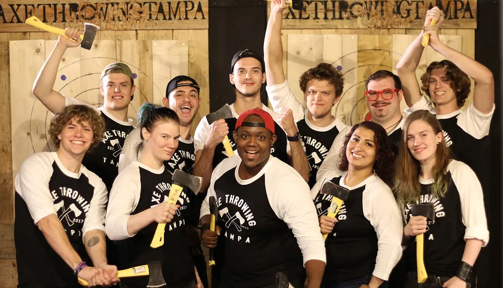 A group of cheerful people wearing matching Axe Throwing Tampa shirts pose with axes in front of a wooden target wall at an axe-throwing venue